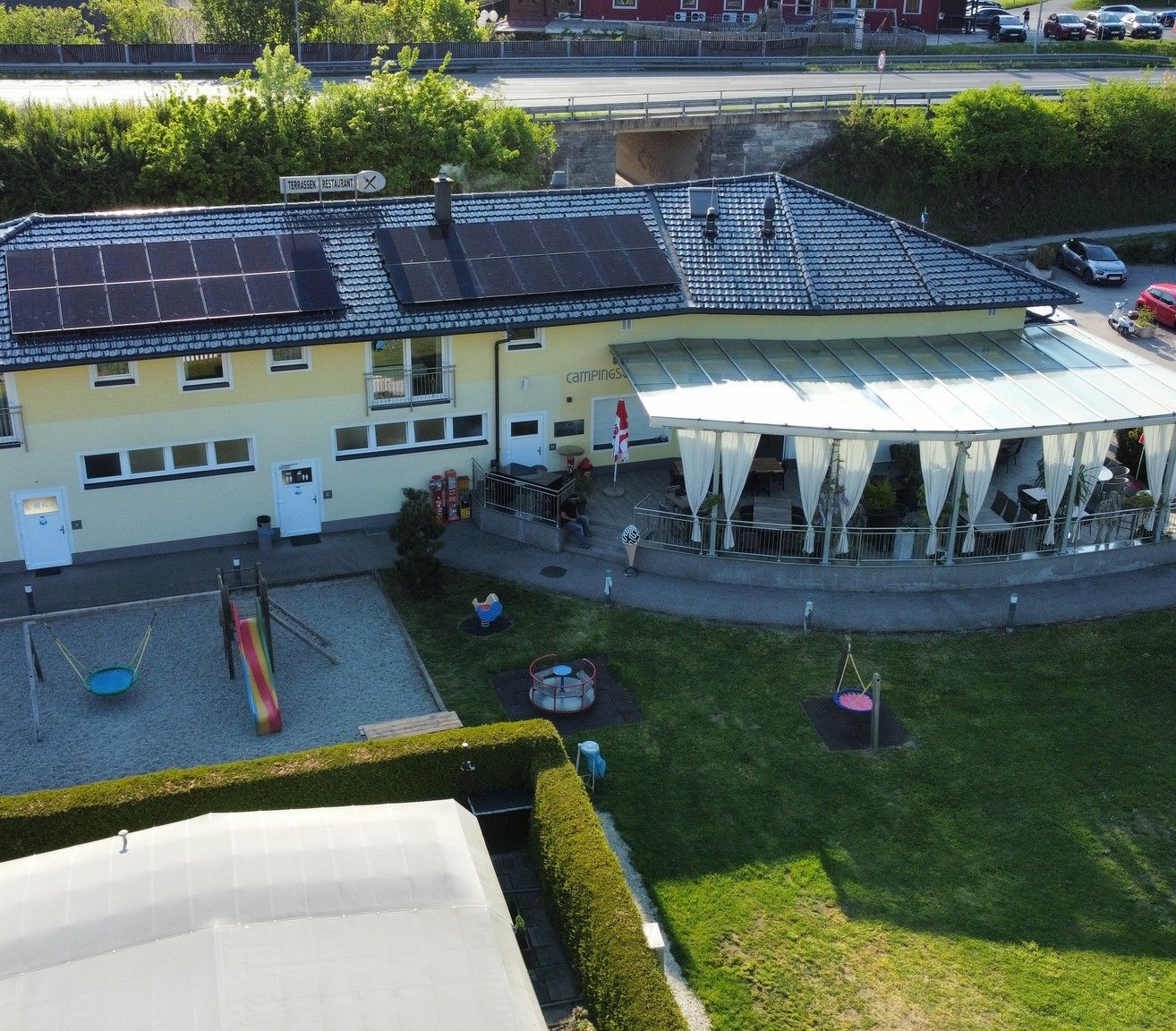 Ein gelbes Gebäude mit Solarmodulen auf dem Dach, einer überdachten Terrasse und einem Spielplatz davor.