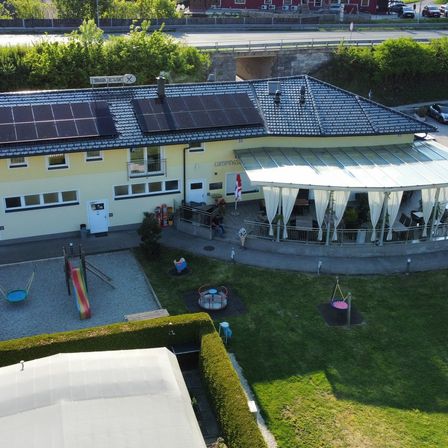Ein gelbes Gebäude mit Solarmodulen auf dem Dach, einer überdachten Terrasse und einem Spielplatz davor.