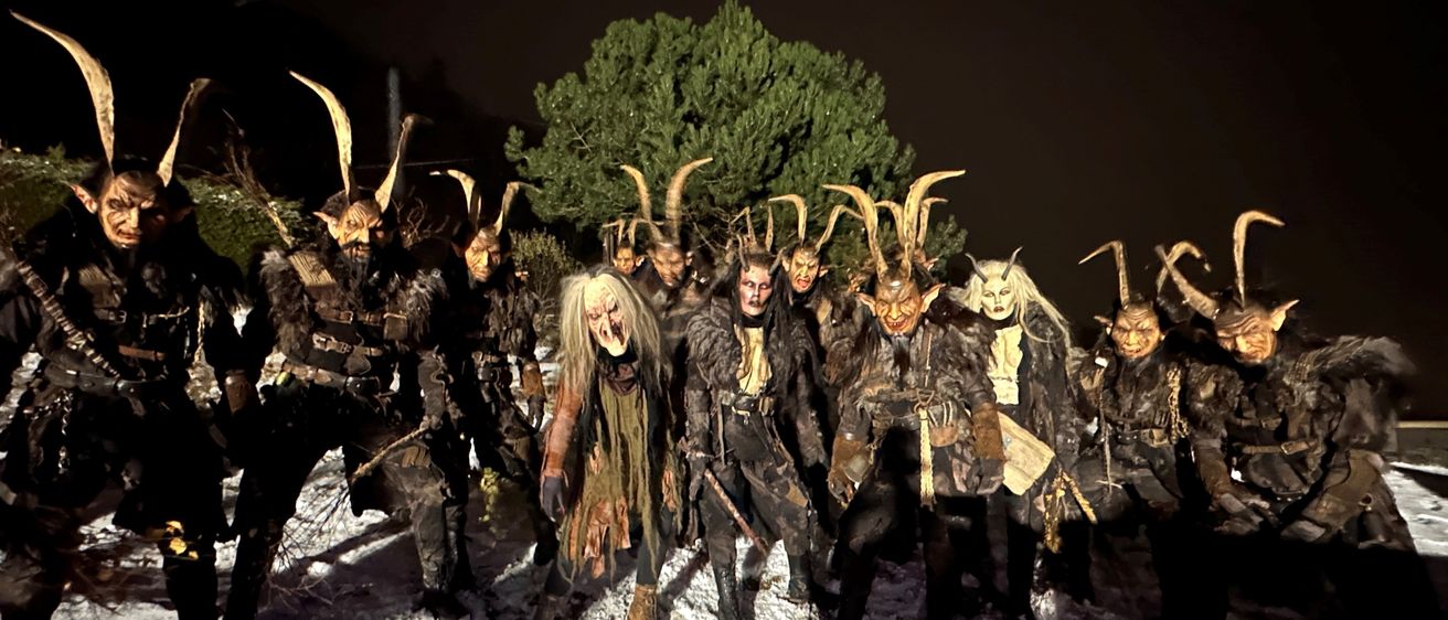 Eine Gruppe von Menschen in Hörnerkostümen steht zusammen im Schnee, eine Person vorne trägt eine weiße Perücke.