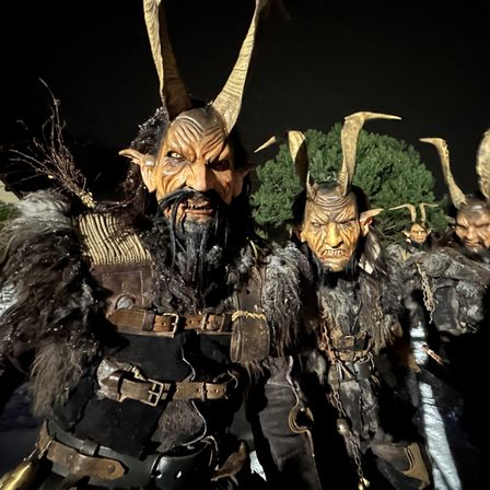 Mehrere Personen in hörnertragenden Dämonenkostümen mit Ketten und Fell scheinen bei einem Festival zu sein. Sie stehen in einer Reihe vor einem dunklen Hintergrund.