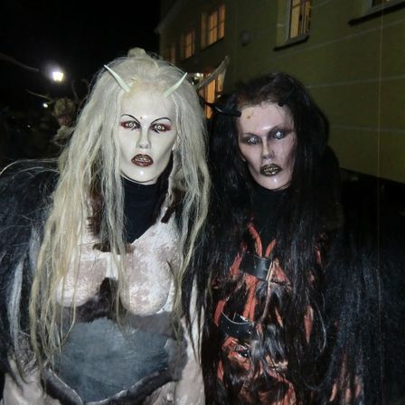 Zwei Personen in aufwendigen Dämonenkostümen stehen bei Nacht zusammen. Die Frau links hat lange blonde Haare mit Hörnern. Die Frau rechts hat lange schwarze Haare und ein rotes Design auf ihrem Kostüm.