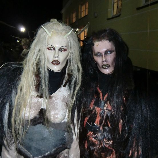 Zwei Personen in aufwendigen Dämonenkostümen stehen bei Nacht zusammen. Die Frau links hat lange blonde Haare mit Hörnern. Die Frau rechts hat lange schwarze Haare und ein rotes Design auf ihrem Kostüm.