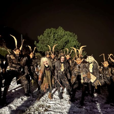 Eine Gruppe von Personen in Hörnerkostümen posiert im Schnee, beleuchtet von einer Taschenlampe, mit einer Kiefer im Hintergrund.