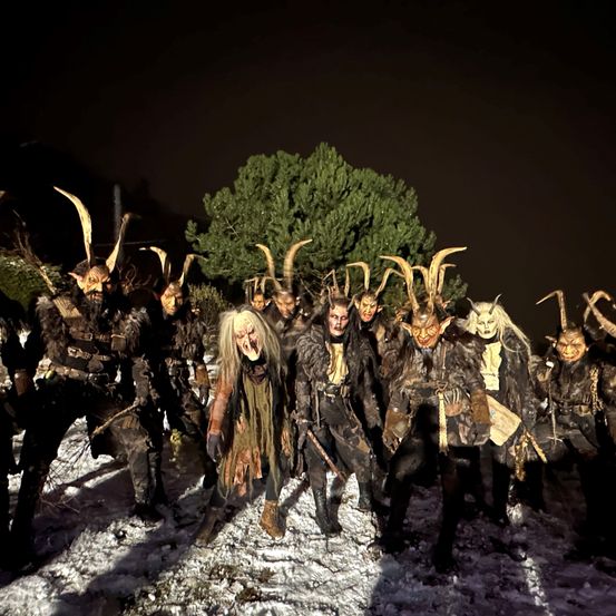 Eine Gruppe von Personen in Hörnerkostümen posiert im Schnee, beleuchtet von einer Taschenlampe, mit einer Kiefer im Hintergrund.