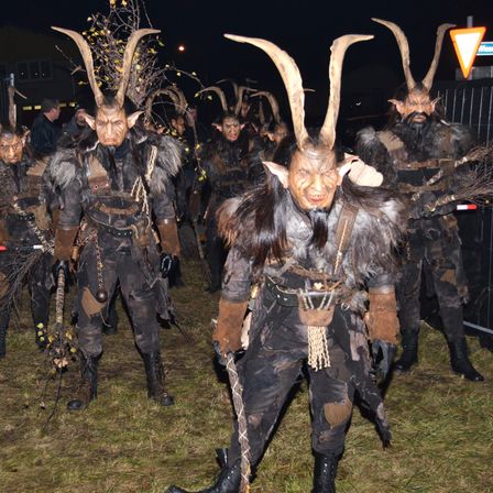 Eine Gruppe von Menschen in Hörnerkostümen läuft bei Nacht auf einem Grasfeld, wahrscheinlich für ein Fest. Die Person vorne hat einen langen Bart und hält einen Stock mit grünen Blättern.