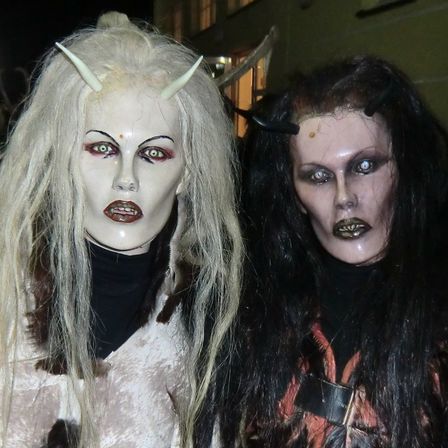 Zwei Frauen in Halloween-Kostümen posieren nachts für ein Foto. Sie tragen aufwendiges Make-up und haben Hörner auf ihren Köpfen. Hinter ihnen steht ein gelbes Gebäude mit Glasfenstern.