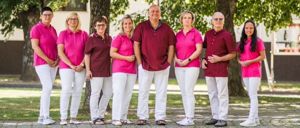 Eine Gruppe von Menschen in passenden rosa und weißen Outfits steht in einem Park, lächelt und posiert für ein Foto. Bäume und Gras sind im Hintergrund zu sehen.