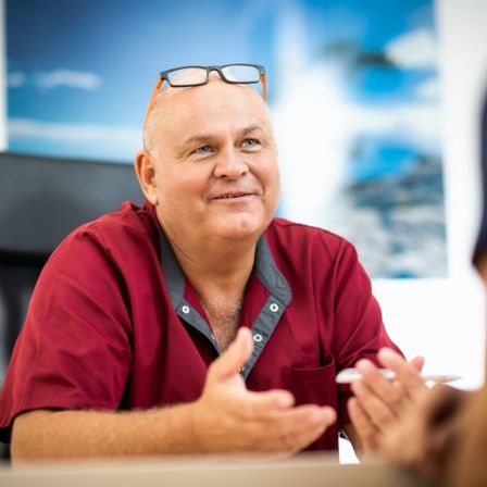 Ein Mann in einem roten Hemd mit einer Brille auf dem Kopf sitzt und lächelt, hält einen Stift und Papier in der Hand. Er scheint in ein Gespräch vertieft zu sein.