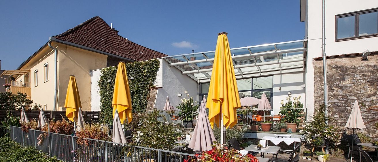 Eine Außencafe-Bereich mit gelben und grauen Sonnenschirmen. Ein Haus mit einem braunen Dach und einem Balkon. Blumentöpfe und Blumen schmücken den Bereich.