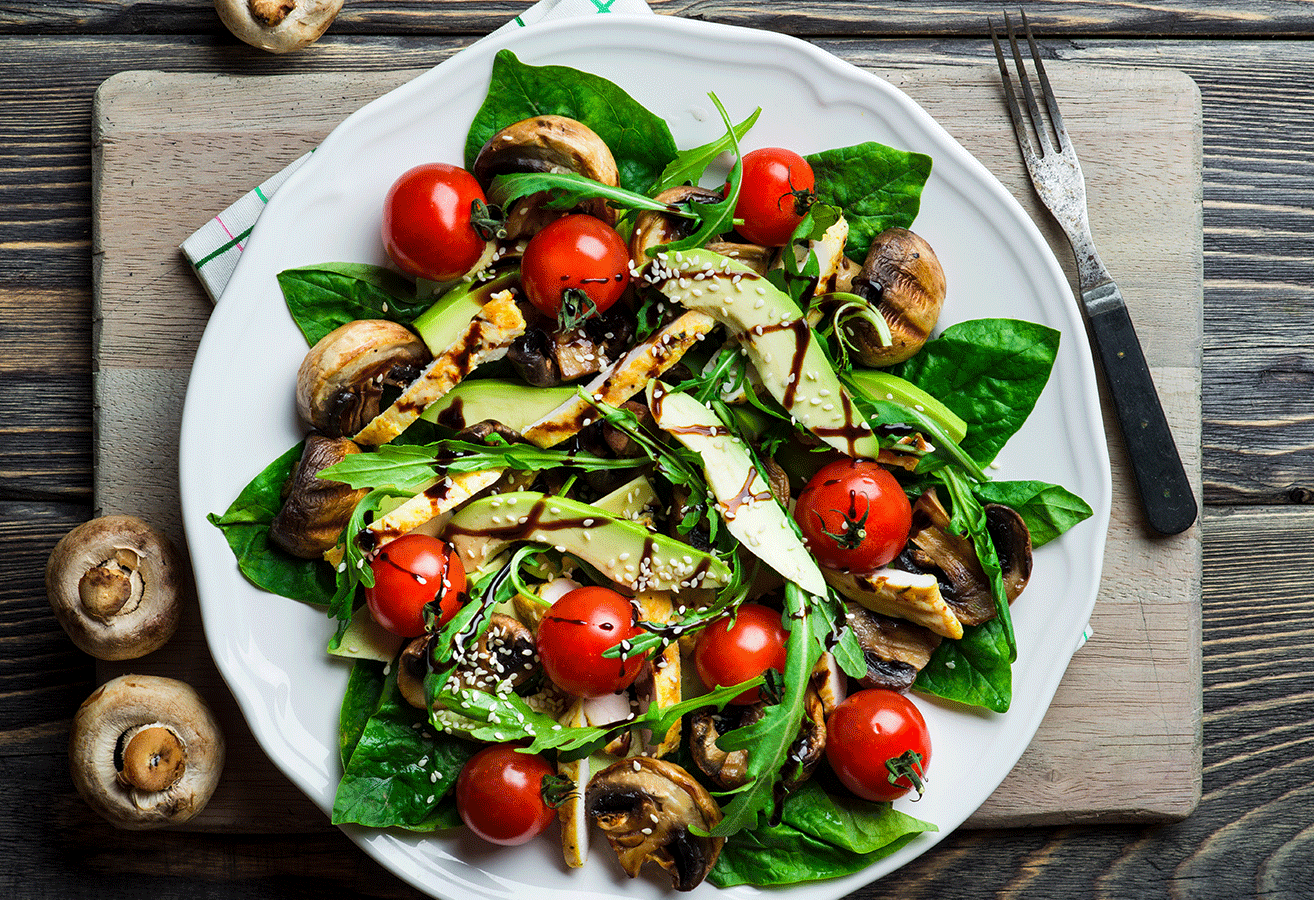 Ein weißer Teller mit Salat aus Spinat, Kirschtomaten, Avocado und Pilzen, garniert mit Sesam und Balsamico-Glasur.