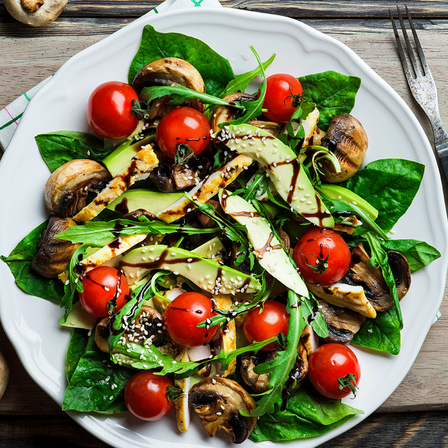 Ein weißer Teller mit Salat aus Spinat, Kirschtomaten, Avocado und Pilzen, garniert mit Sesam und Balsamico-Glasur.