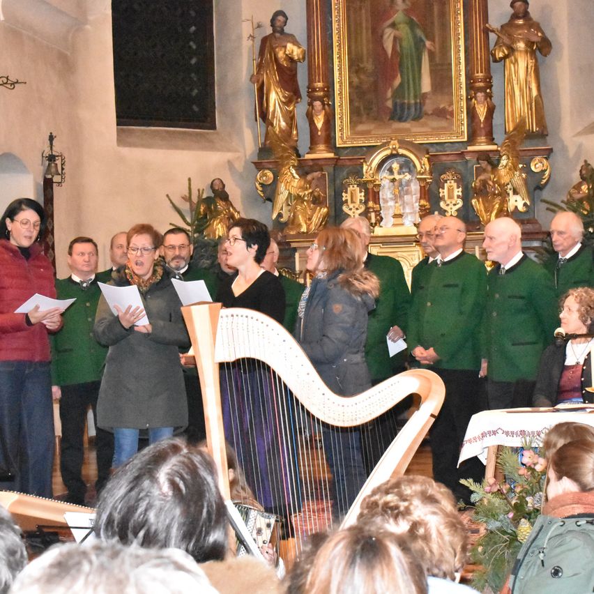 Ein Chor singt in einer Kirche mit einem goldenen Altarbild und Statuen. Die Chormitglieder stehen hinter einer Harfe und singen, während sie Notenblätter halten. Das Publikum sitzt vor ihnen.