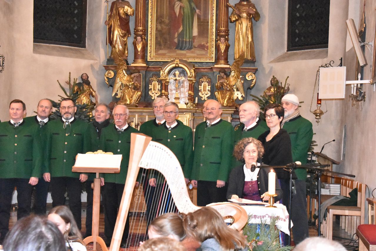 Eine Gruppe von Männern und Frauen in grünen Uniformen steht in einer Kirche. Eine Frau liest ein Buch und eine Kerze steht auf dem Schreibtisch. Eine Harfe ist auf einem Ständer platziert und einige Menschen stehen hinter ihnen. Dahinter befindet sich eine goldene Statue einer Person.