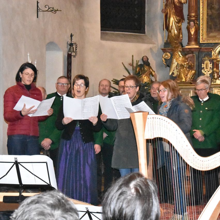 Ein Chor in einer Kirche singt und hält Notenblätter. Eine Frau spielt vor ihnen die Harfe. Mehrere Menschen stehen hinter ihnen.