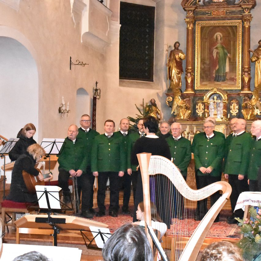 Ein Chor in grünen Kleidern steht in einer Kirche mit einem großen goldenen Altarbild und einer Harfenistin. Eine Frau spielt Gitarre. Das Publikum sitzt vor ihnen.