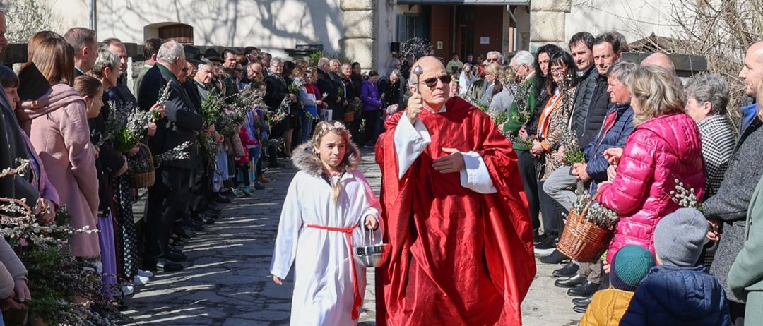 Ein Priester in einem roten Gewand geht vor einer Menschenmenge, die Blumenzweige hält.