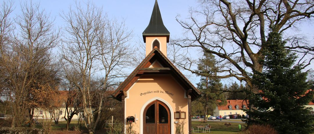 Eine kleine Kapelle mit einem Turm, umgeben von Bäumen und Gras, hat eine hölzerne Tür und ein Schild mit der Aufschrift 'Begrüßer seid ihr, Maria'. Ein Weg führt zum Eingang.