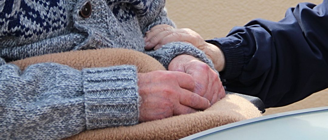 Zwei ältere Personen halten Händchen, einer trägt einen gestrickten Pullover, und sitzen auf einer mit Decken bedeckten Couch.
