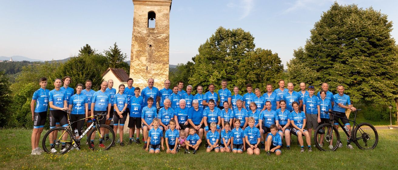 Eine Gruppe von Radfahrern in blauen Trikots posiert für ein Foto vor einem historischen Turm und üppigem Grün unter einem klaren Himmel.