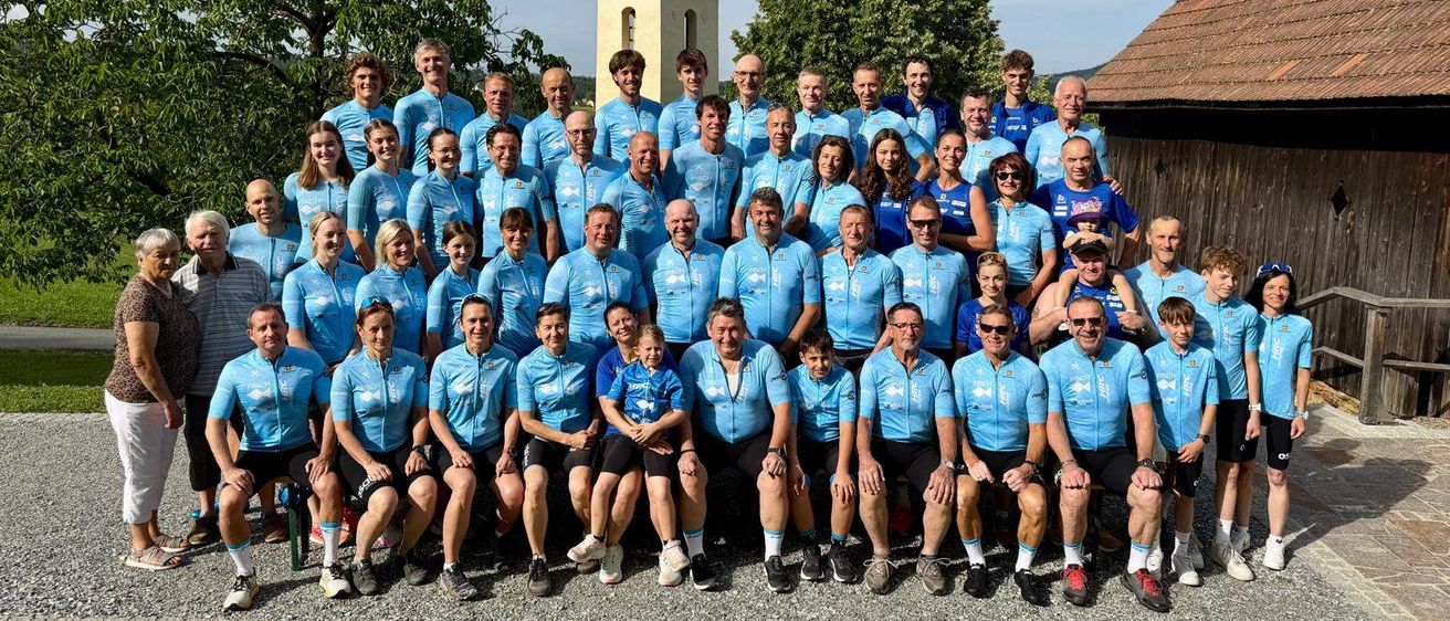 Eine Gruppe von Radfahrern in blauen Trikots posiert für ein Foto vor einer Kirche und Bäumen.