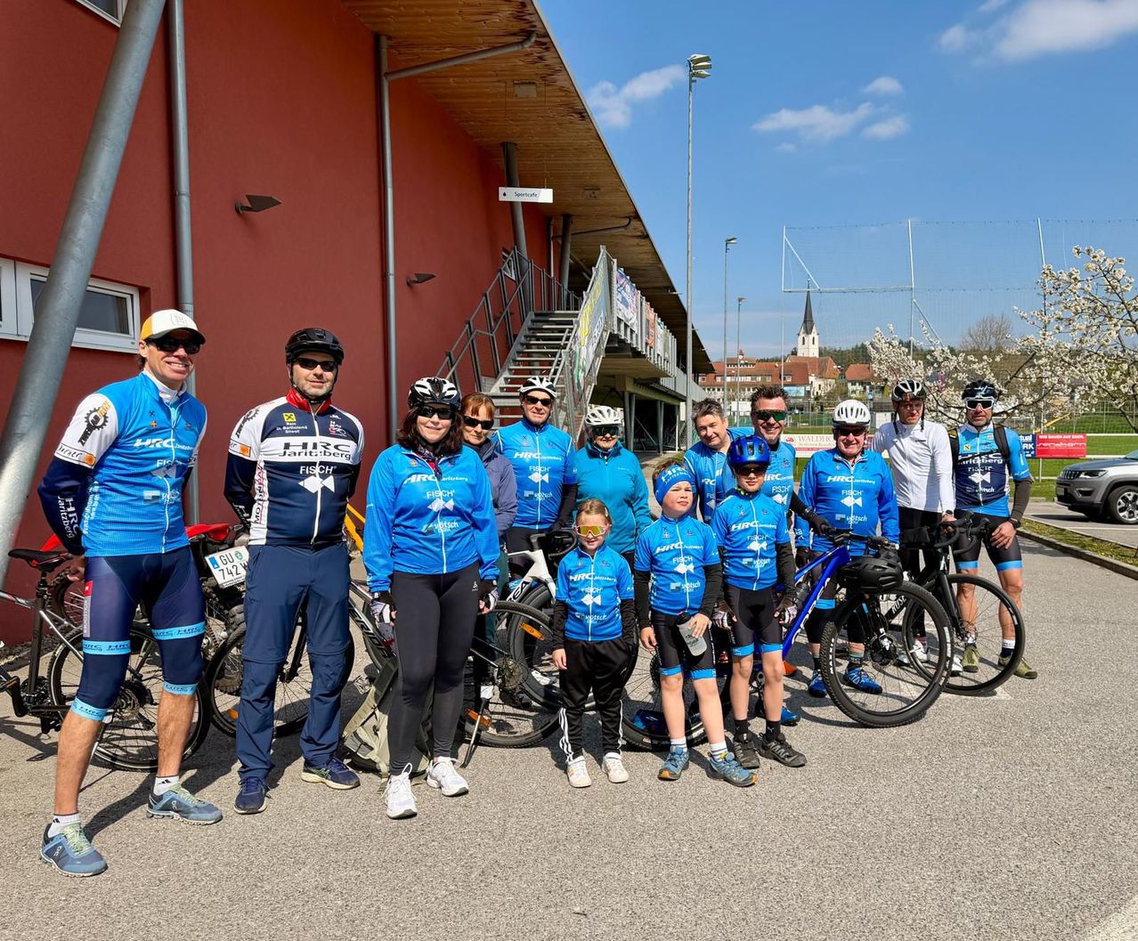 A group of cyclists in matching blue uniforms pose for a photo in front of a building with a red wall, near parked bicycles.