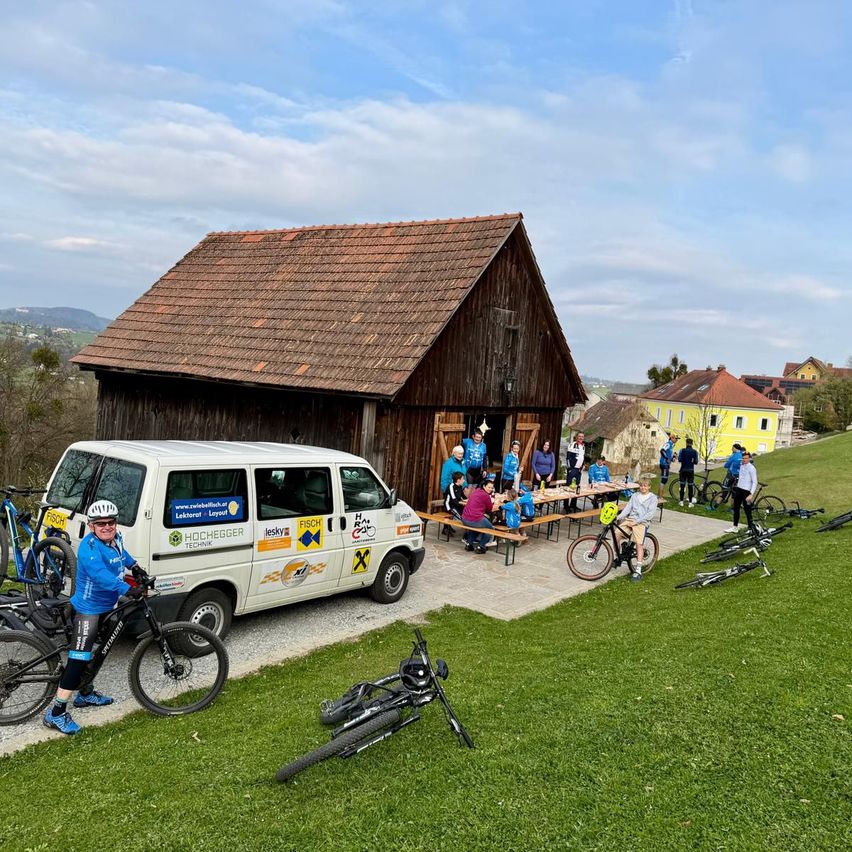 A group of cyclists gather outside a barn with a van nearby. Some are sitting at a picnic table, others are standing around. The sky is cloudy.