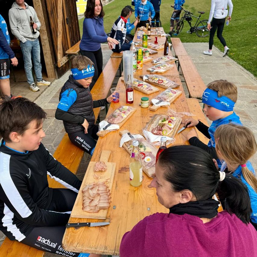 A group of cyclists and their families are sitting at a long table, enjoying a meal with various foods and drinks on it.