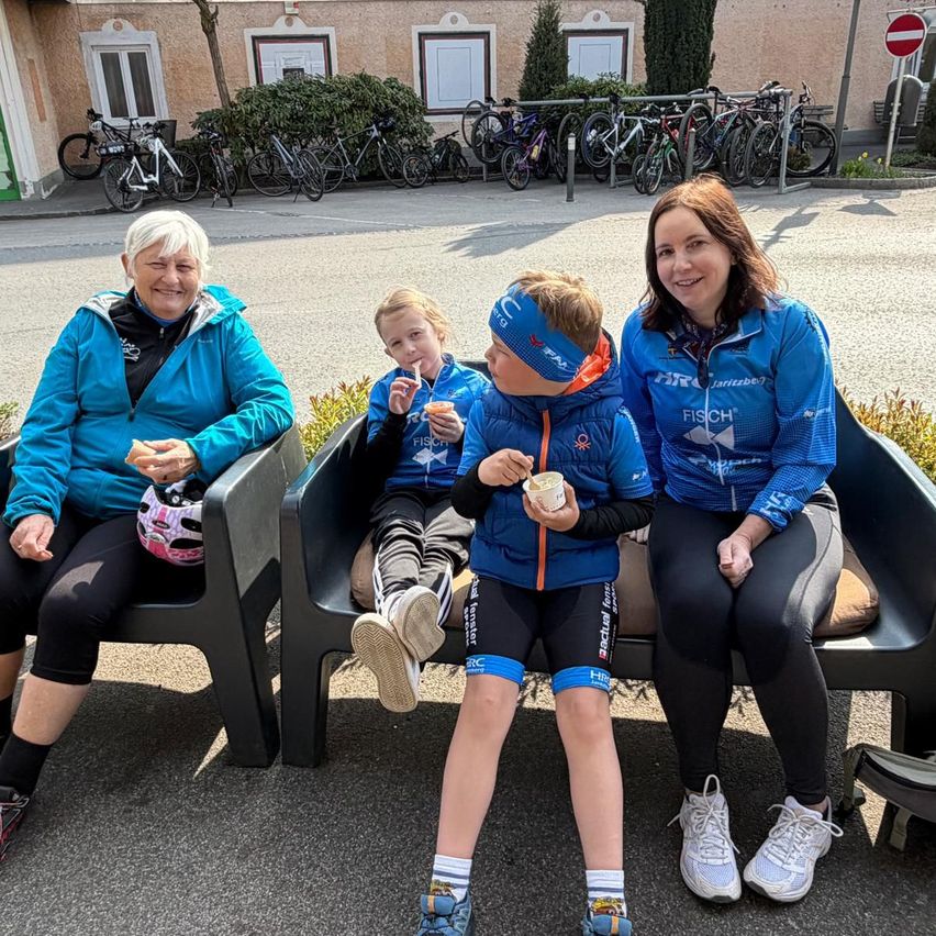 Three people sit on a bench, smiling. A woman holds a helmet. Two children hold ice cream, one wears a headband. Bikes and a building are in the background.