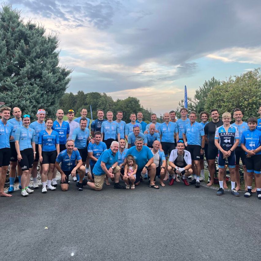 Eine Gruppe von Radfahrern in blauen Trikots posiert für ein Foto auf einer Straße mit Bäumen und einem bewölkten Himmel im Hintergrund.