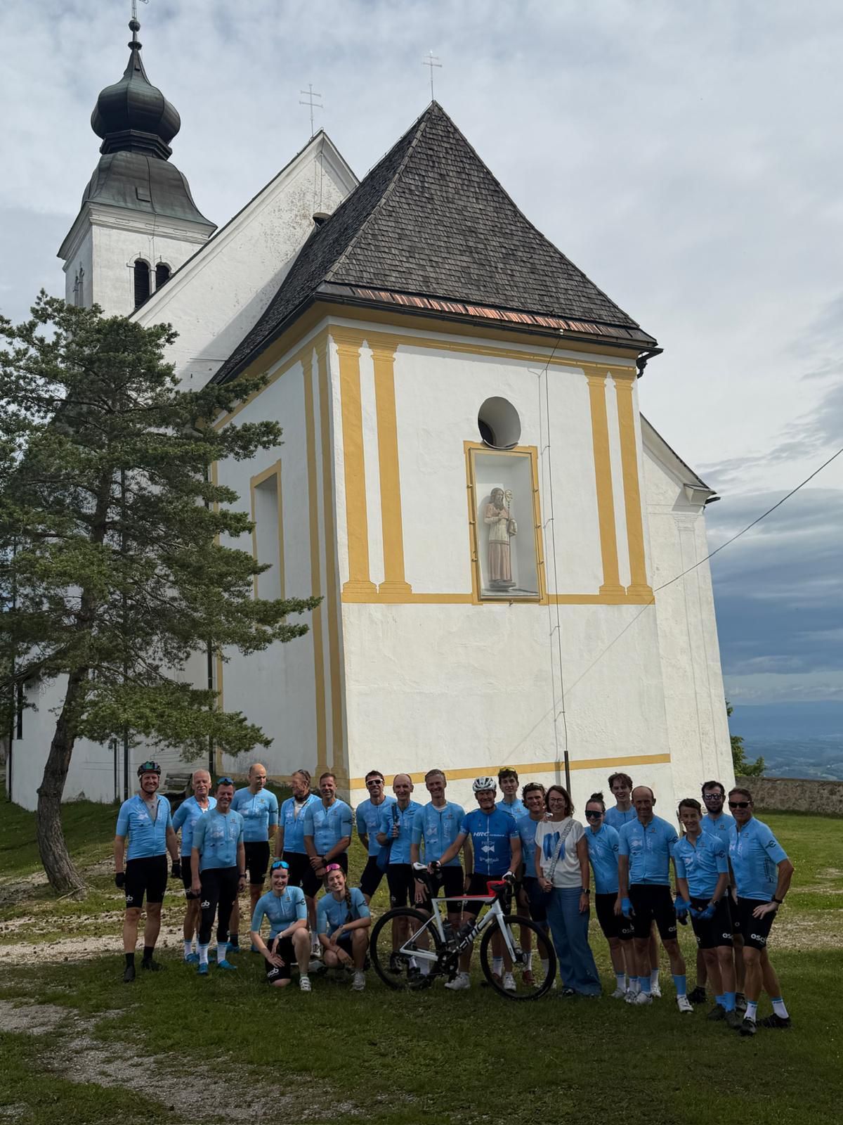 Eine Gruppe von Radfahrern in blauen Trikots posiert für ein Foto vor einer Kirche mit gelben Akzenten und einer Statue eines Heiligen.