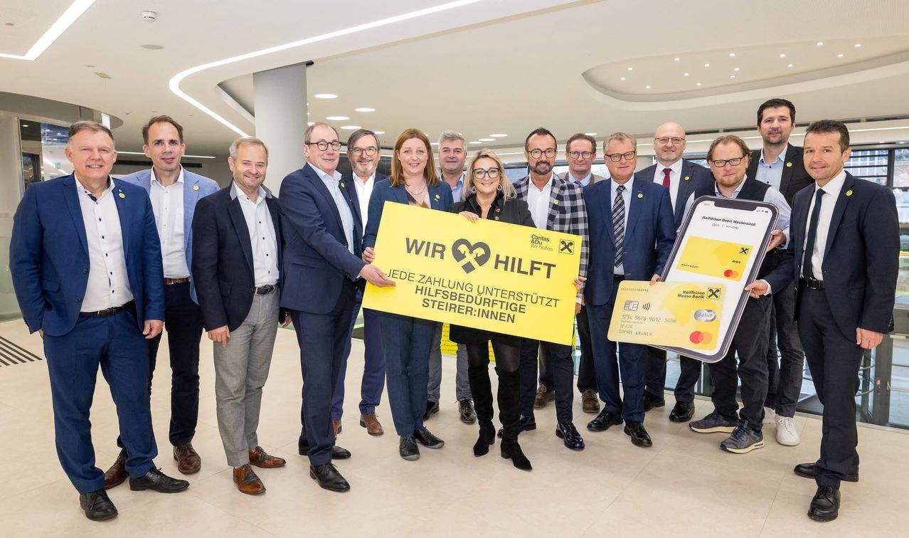 Eine Gruppe formell gekleideter Personen steht in einer modernen Lobby und hält ein gelbes Schild mit der Aufschrift 'Wir hilft'. Sie lächeln und posieren für ein Foto. Einige halten eine gelbe Karte.