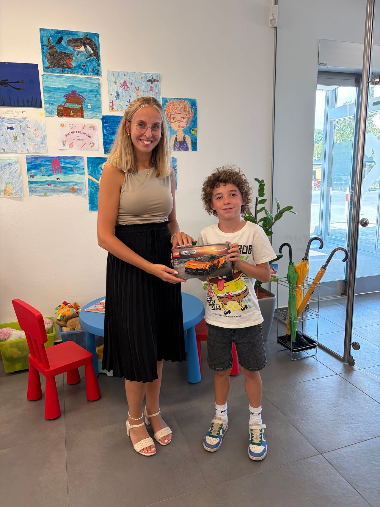 Eine Frau und ein Junge stehen in einem Raum mit farbenfrohen Gemälden an der Wand und halten ein Spielzeugauto. Der Raum hat einen blauen Tisch, einen roten Stuhl und eine Pflanze.