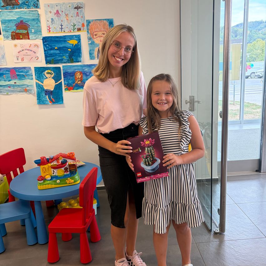 Eine Frau und ein junges Mädchen stehen in einem Raum mit einem Tisch, Stühlen und einer Glastür. Das Mädchen hält eine LEGO-Box mit einem Kaktus. Hinter ihnen zeigt eine Wand Kinderkunst.