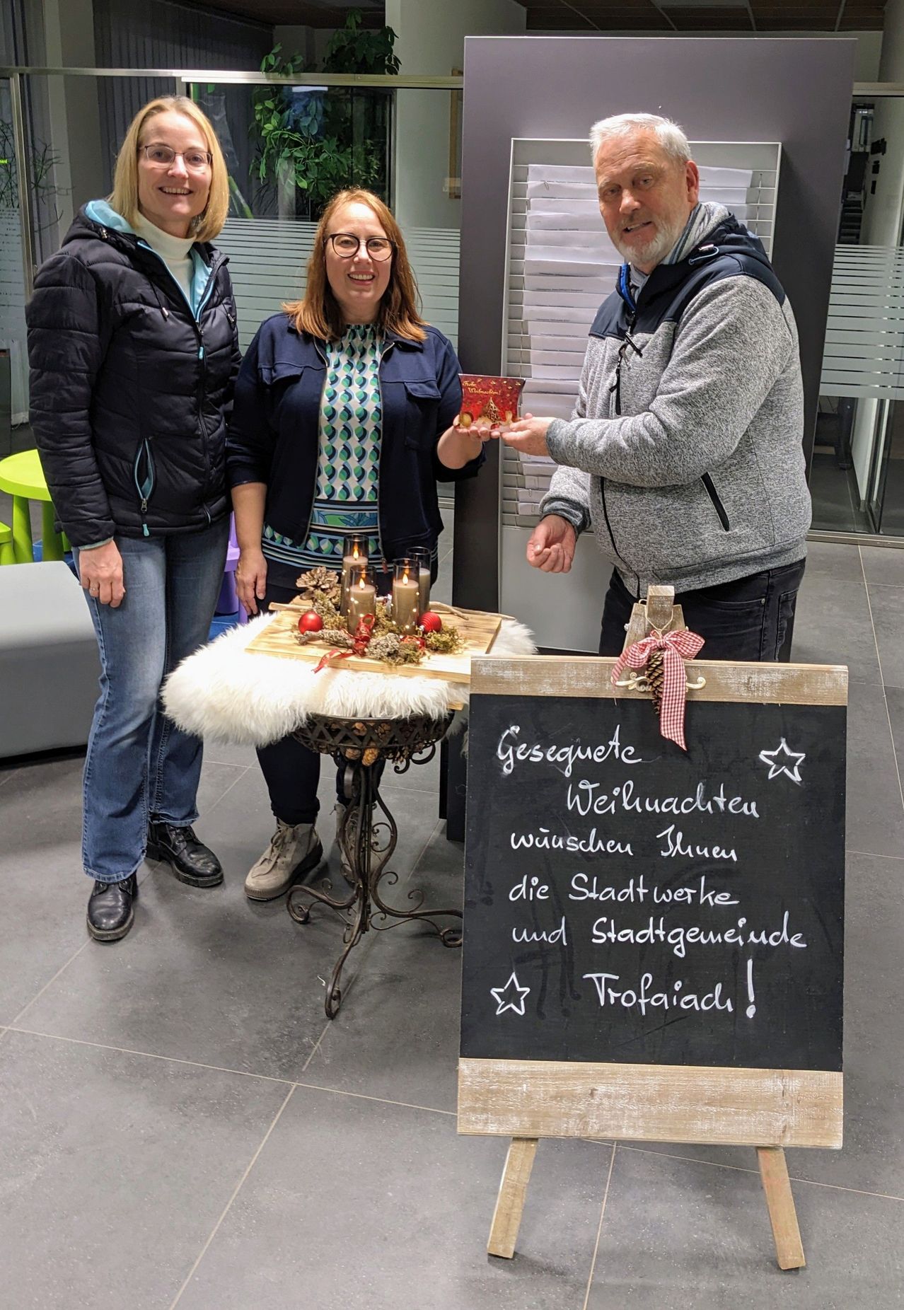 Drei Personen stehen zusammen bei einem festlich gedeckten Tisch mit Kerzen und einer Tafel, auf der 'Gesegnete Weihnachten' steht. Ein Mann überreicht einer Frau ein Geschenk.
