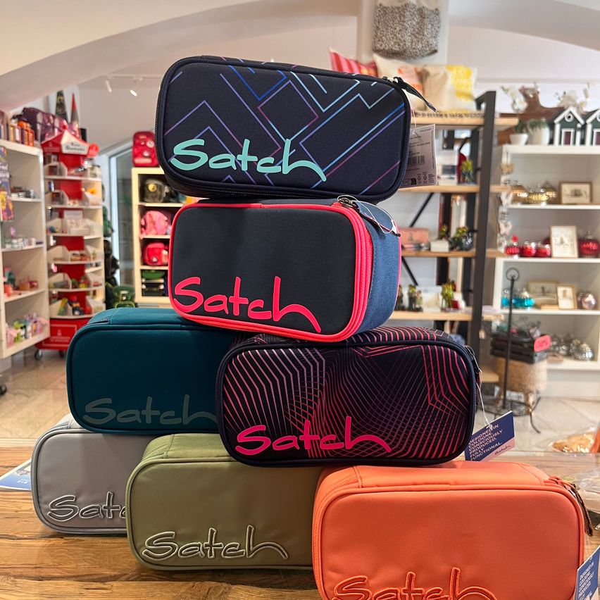 A stack of colorful Satch pencil cases with the brand name prominently displayed, arranged on a wooden table in a store.