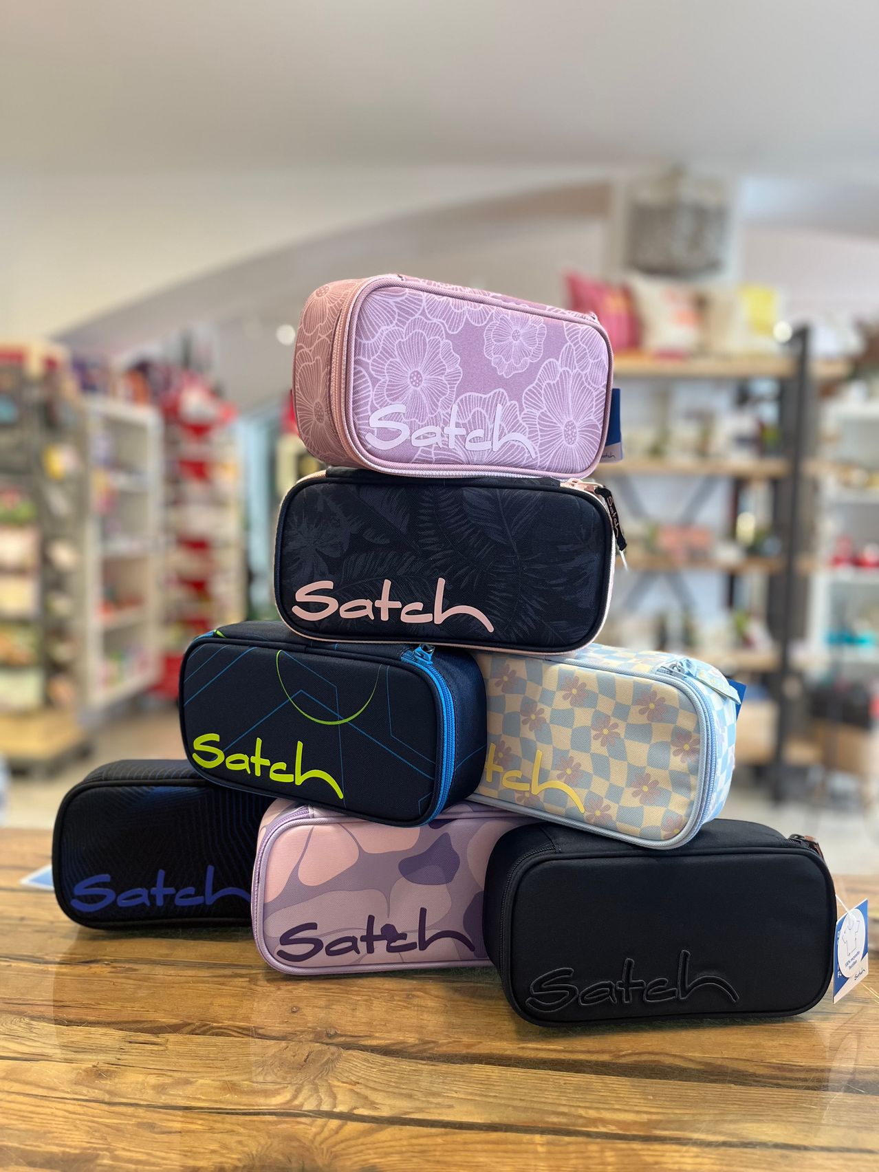 A stack of colorful Satch pencil cases on a wooden table in a store.