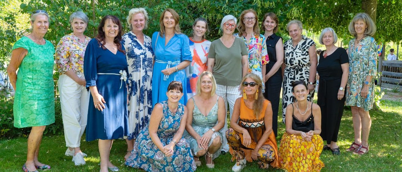 Eine Gruppe von Frauen steht und sitzt auf einem Rasen unter einem großen Baum, gekleidet in verschiedene Sommeroutfits. Sie lächeln und posieren für ein Foto.