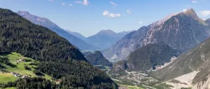 Ein Panoramablick auf eine bergige Region mit dichten Wäldern, einem kleinen Ort im Tal und einer Burg auf einem Hügel unter einem klaren blauen Himmel.