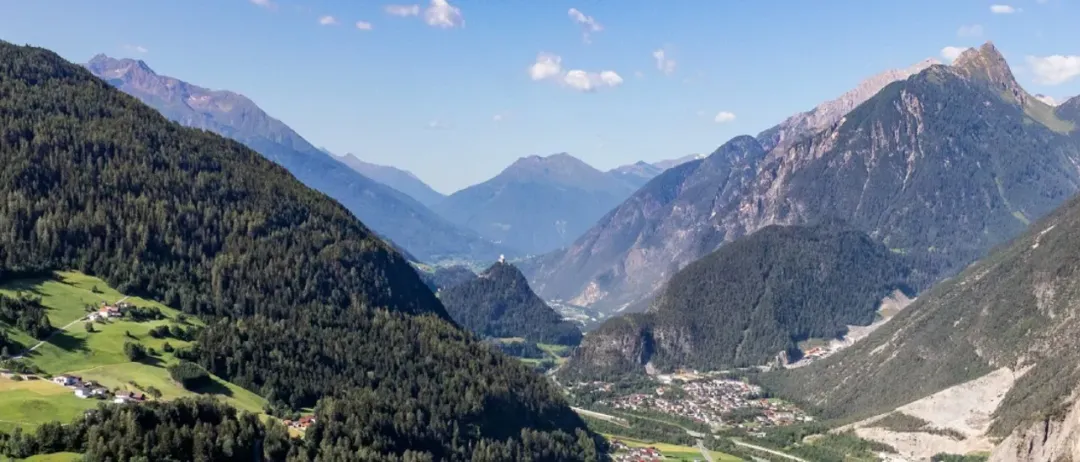 Ein Panoramablick auf eine bergige Region mit dichten Wäldern, einem kleinen Ort im Tal und einer Burg auf einem Hügel unter einem klaren blauen Himmel.
