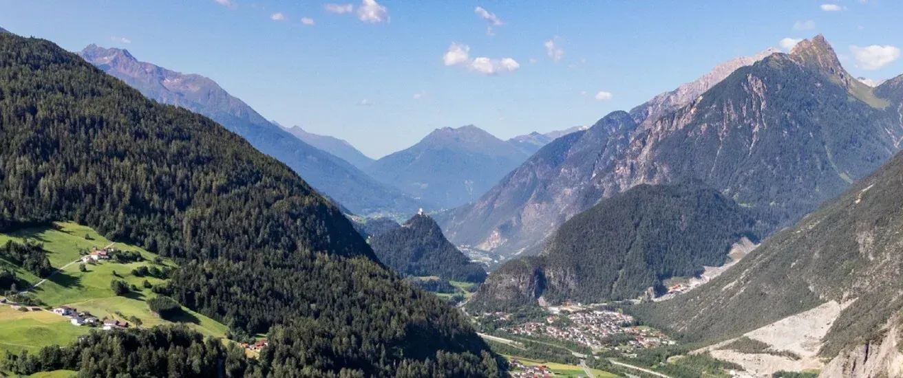 Ein Panoramablick auf eine bergige Region mit dichten Wäldern, einem kleinen Ort im Tal und einer Burg auf einem Hügel unter einem klaren blauen Himmel.