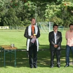 Drei Männer stehen in einem Park mit einem Fußballfeld im Hintergrund. Ein Mann hält ein Buch und einen Tennisball.