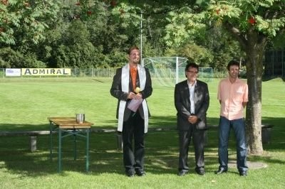 Drei Männer stehen in einem Park mit einem Fußballfeld im Hintergrund. Ein Mann hält ein Buch und einen Tennisball.