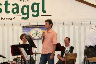 Ein Mann steht auf der Bühne mit einem Mikrofon und spricht das Publikum an. Hinter ihm spielen Musiker Instrumente, und ein Banner mit Text ist sichtbar.