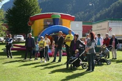 Eine Gruppe von Menschen, darunter Erwachsene und Kinder, versammelt sich um eine aufblasbare Hüpfburg in einem Park mit Bergen im Hintergrund.