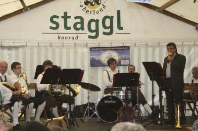 Eine Band tritt auf der Bühne unter einem Zelt auf, ein Mann in Anzug singt in ein Mikrofon. Die Band besteht aus Tuba-, Schlagzeug- und Trompetenspielern. Das Publikum sitzt vor ihnen.