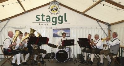 Eine Blechbläsergruppe spielt auf der Bühne unter einem weißen Zelt mit dem Wort 'Stagggl' im Hintergrund. Die Musiker spielen verschiedene Instrumente, darunter Trompeten, Posaunen und Tuben.