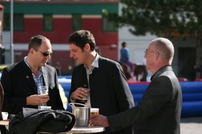 Drei Männer in Anzügen stehen um einen Tisch herum, einer hält ein Glas Bier und ein anderer einen Eimer Bier. Sie führen ein Gespräch. Hinter ihnen befindet sich eine blaue aufblasbare Struktur und ein Gebäude.