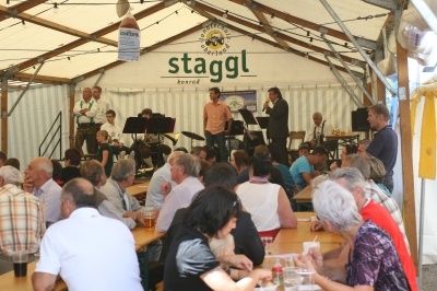Eine Gruppe von Menschen versammelt sich in einem Zelt für eine musikalische Aufführung. Eine Band spielt auf der Bühne, und das Publikum sitzt an Tischen und genießt Getränke und Essen.