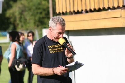 Ein Mann mit grauem Haar hält ein Mikrofon und ein Papier in der Hand und spricht möglicherweise bei einer Veranstaltung. Er trägt ein schwarzes T-Shirt mit gelbem Text. Hinter ihm stehen mehrere Menschen und unterhalten sich.