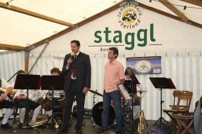 Zwei Männer stehen auf einer Bühne mit einer Band dahinter, einer hält ein Mikrofon und der andere ein Notenständer. Die Bühne ist in einem Zelt aufgebaut mit einem großen Schild, das 'staggl' besagt.